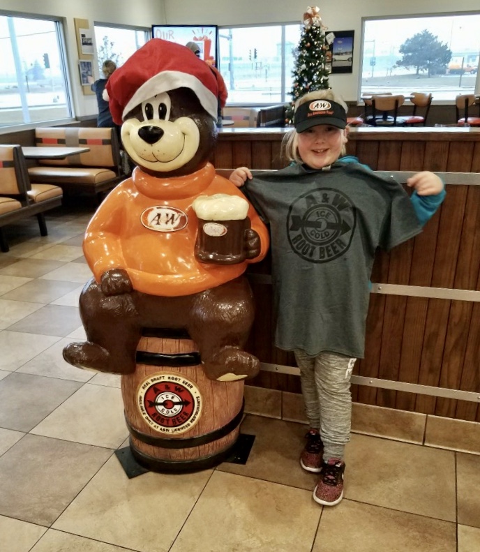 Photo of Abby holding a gray A&W t-shirt next to a statue of Rooty the Great Root Bear